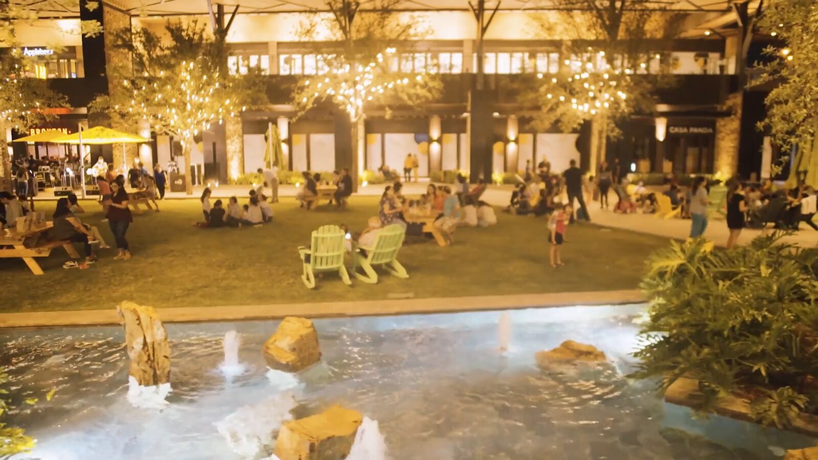 Pueblo Serena de noche: fuente iluminada, sillas y series de luces entre los locales